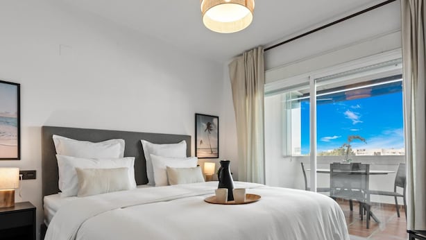 Una habitación de hotel con una cama grande, una mesita de noche, una ventana con vistas y un balcón con mesa y sillas.