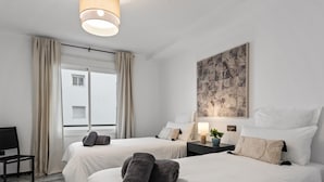 Habitación de hotel con dos camas, un escritorio, una silla, un ventanal con cortinas y un cuadro en la pared.