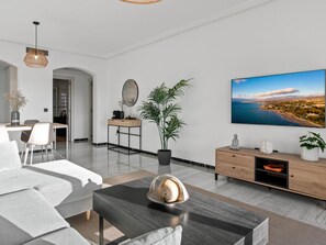 Ein modernes Wohnzimmer mit einem großen Flachbildfernseher, einem Couchtisch mit Dekoration, einer Topfpflanze und einem schlichten Arbeitsbereich.
