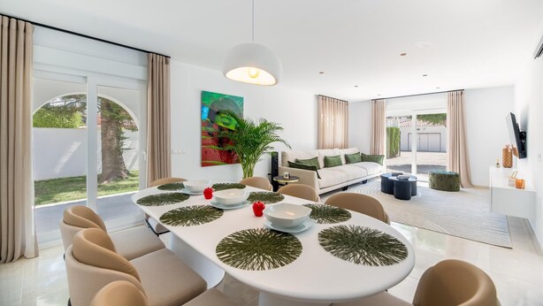 Ein modernes Wohnzimmer mit einer Couch, einem Esstisch und Stühlen. Ein großes Fenster mit Vorhängen, eine Pflanze und ein Gemälde an der Wand.