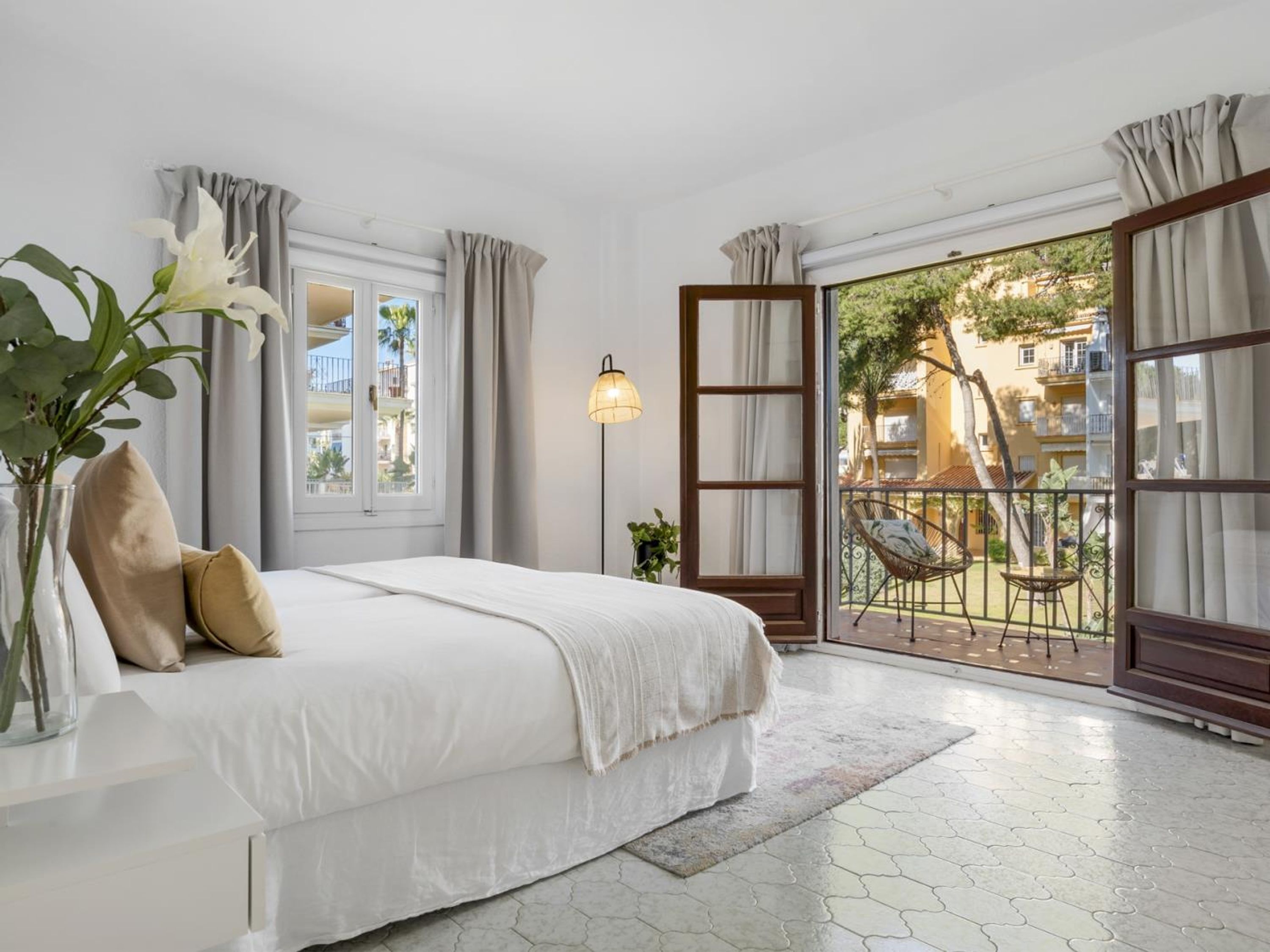 A bedroom with a bed, a bedside plant, a lamp, and a view of the outside balcony.