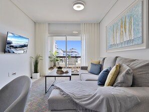 A modern living room with a sofa, a flat-screen TV, a coffee table, and a balcony with a view of buildings. - 1 Dormitorio en Medina Garden (Marbella)