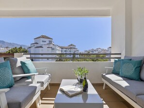 A balcony with a sofa, armchairs, and a table, overlooking a coastal town with white buildings and mountains in the distance. - 1 Dormitorio en Medina Garden (Marbella)