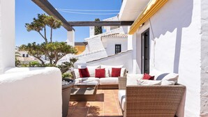 Une terrasse ensoleillée avec un mur blanc, un canapé en osier et une petite table agrémentée de coussins rouges.