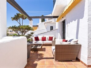 A sunlit patio with a white wall, a wicker sofa, and a small table with red cushions. - 3 Bedroom Townhouse Aldea Blanca (Marbella)