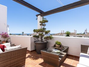 A balcony with wicker furniture, a potted tree, and a view of the sea. - 3 Bedroom Townhouse Aldea Blanca (Marbella)