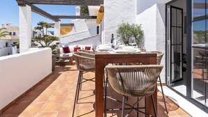 Une terrasse extérieure avec une table en bois et des chaises en osier, un mur blanc et une porte vitrée donnant sur un balcon.