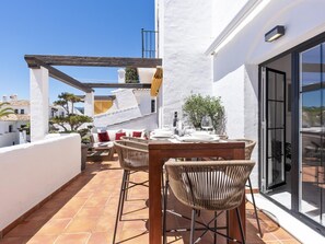 An outdoor patio with a wooden table and wicker chairs, a white wall, and a glass door leading to a balcony. - 3 Bedroom Townhouse Aldea Blanca (Marbella)