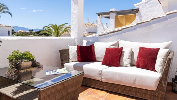 Un balcon avec un canapé blanc, des coussins rouges, une table basse en verre et une plante en pot.
