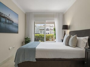 Ein Hotelzimmer mit Bett, Nachttisch, Lampe, einem Gemälde und Blick auf ein Gebäude durch ein Fenster.