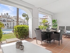 Eine sonnendurchflutete Terrasse mit Esstisch und Stühlen, einer weißen Wand und Blick auf einen Swimmingpool und Gebäude.