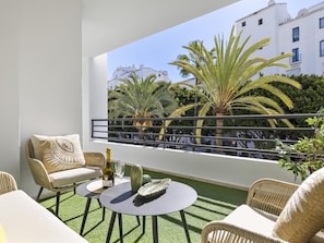 A balcony with a view of palm trees and buildings, furnished with wicker chairs and a small table with a bottle. - Terrazas DE Banus 4 12 (Marbella)