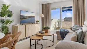 Uma sala de estar moderna com televisão, sofá, mesa de centro e vista para a água através de grandes janelas.