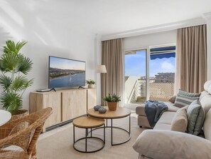 Ein modernes Wohnzimmer mit Fernseher, einer Couch, einem Couchtisch und Blick auf ein Gewässer durch große Fenster.