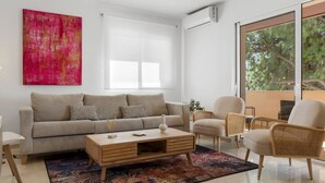 En moderne stue med sofa, sofabord og stole, et stort vindue med udsigt til træer, og et farverigt abstrakt billede på væggen.