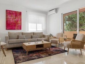 Ein modernes Wohnzimmer mit einer Couch, einem Couchtisch und Sesseln, ein großes Fenster mit Blick auf Bäume und ein leuchtendes abstraktes Gemälde an der Wand.