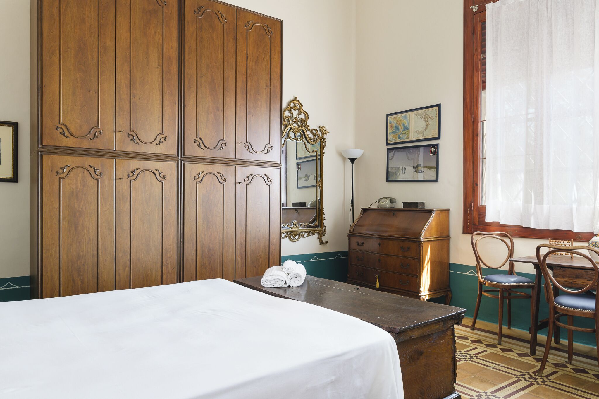 Ein Hotelzimmer mit großem Holzschrank, Schreibtisch mit Telefon, zwei Stühlen und einem Bett mit weißer Bettdecke.