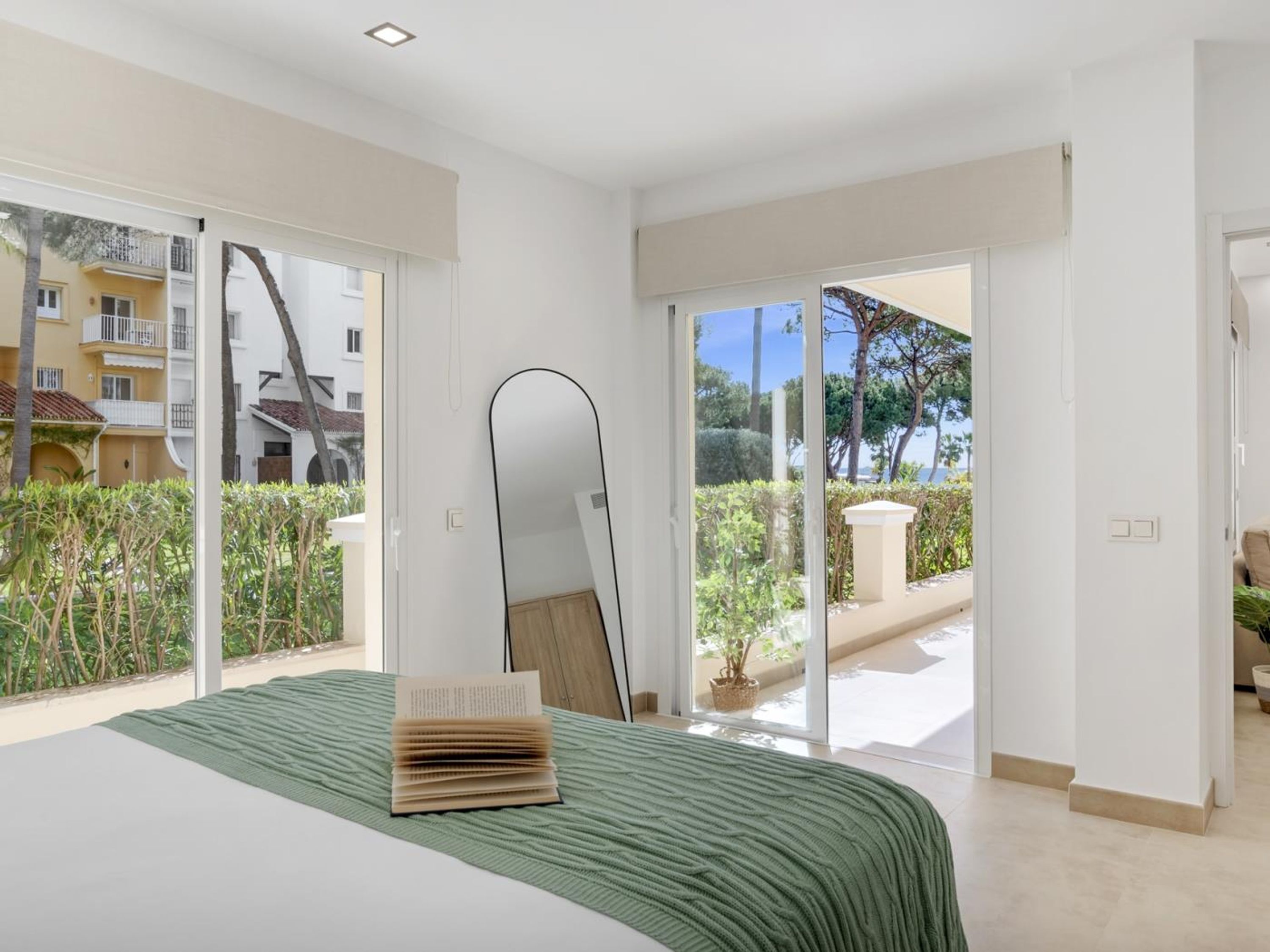 Un dormitorio con cama, un espejo grande y vistas a un balcón con plantas y un edificio.