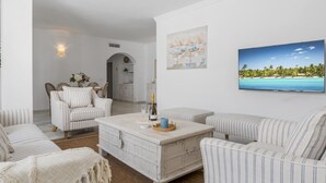 Un soggiorno con un divano bianco, una poltrona a righe e un tavolino da caffè con una bottiglia e dei bicchieri. Al muro, un quadro raffigura una spiaggia.