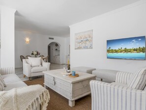 Ein Wohnzimmer mit einem weißen Sofa, einem gestreiften Sessel und einem Couchtisch mit einer Flasche und Gläsern. An der Wand hängt ein Gemälde, das eine Strandansicht zeigt.