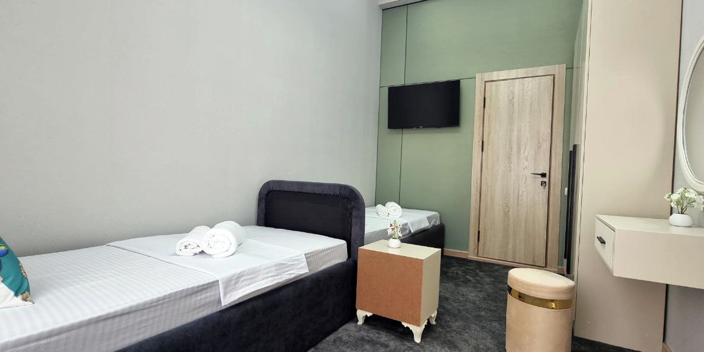 A hotel room with two single beds, a small bedside table, a trash bin, a mirror, and a television on the wall.
