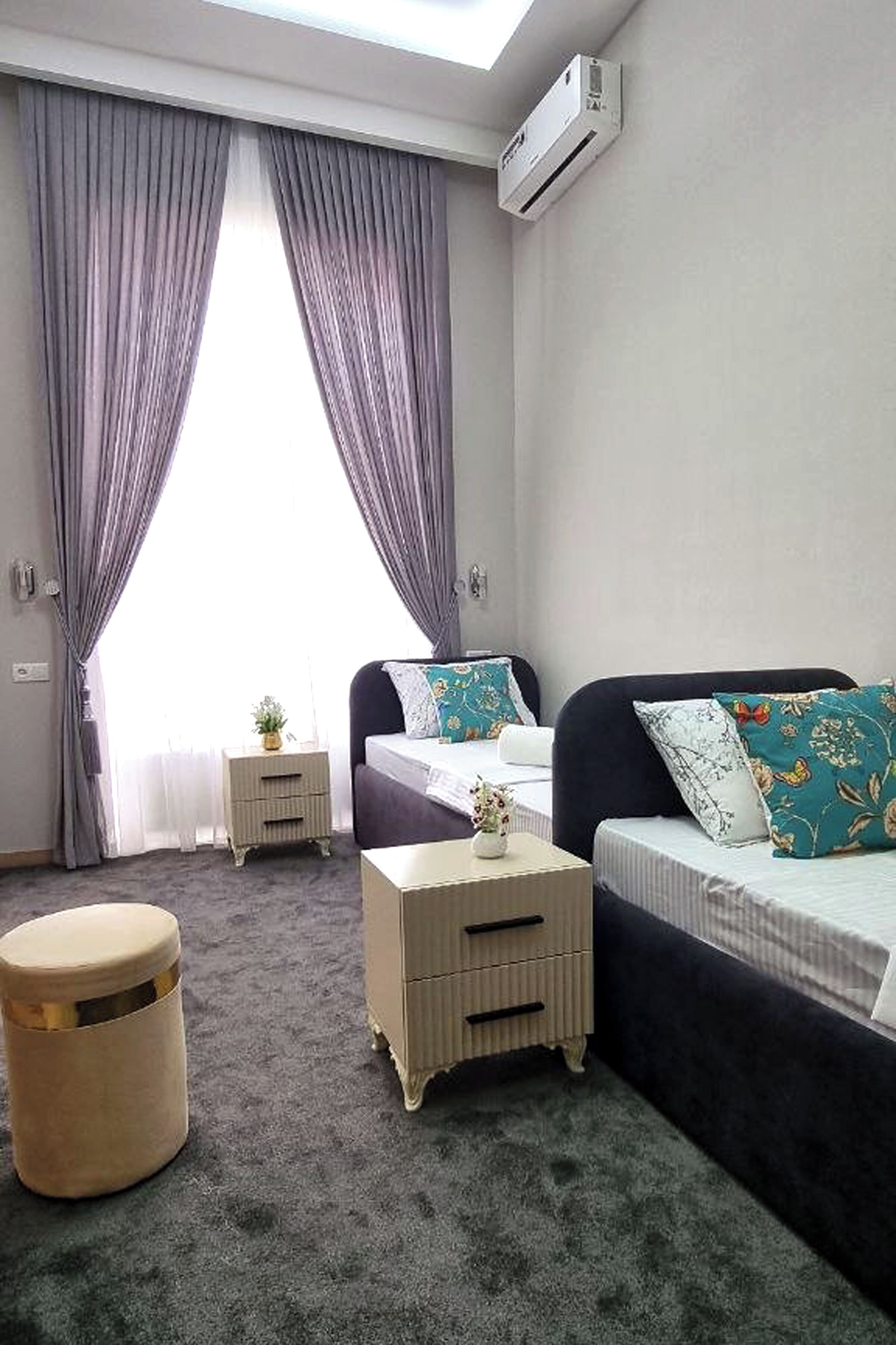 A bedroom with a bed, a bedside table, a small stool, and a window with curtains.