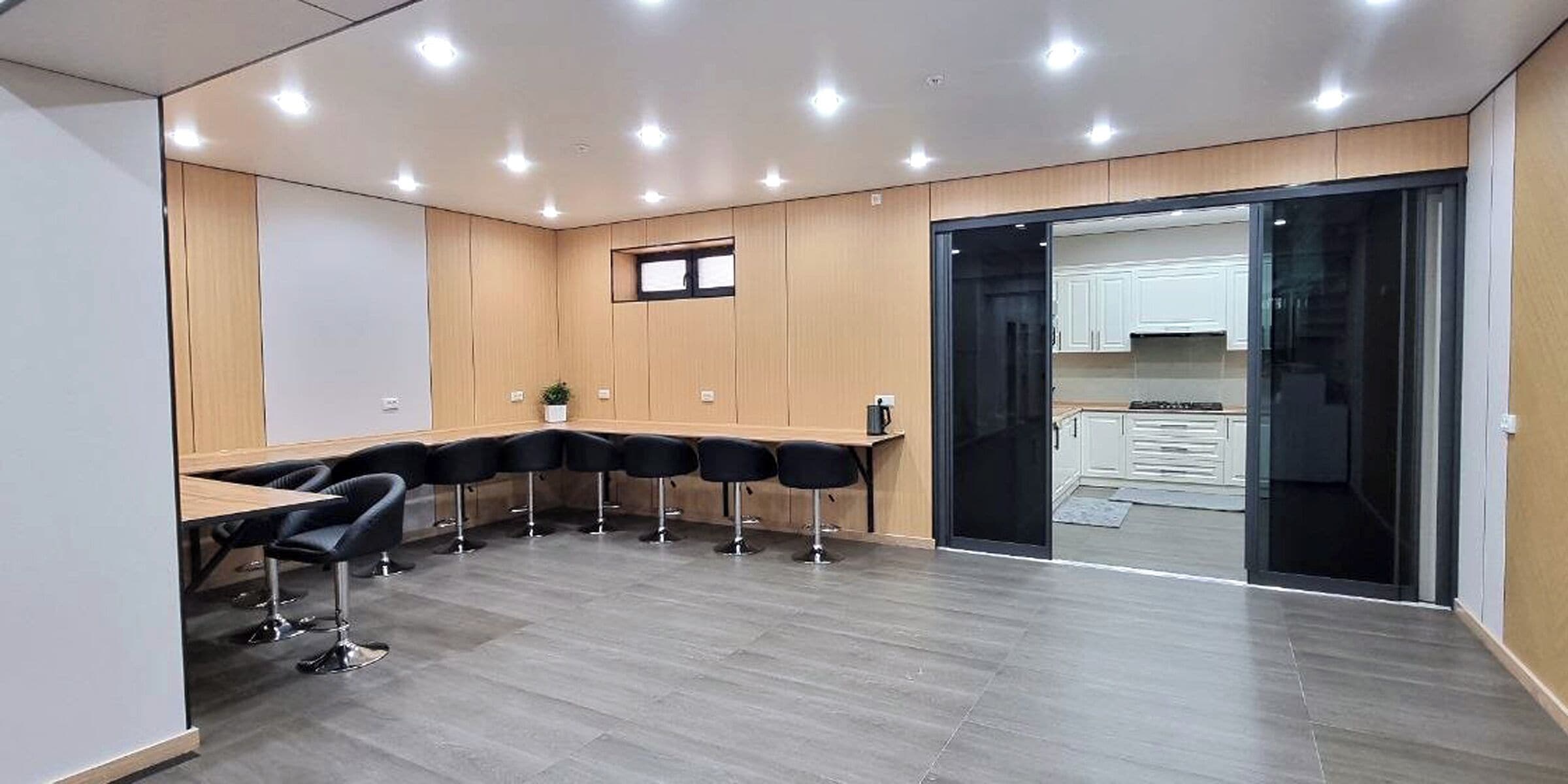 A conference room with a long table and chairs, a small window, and a kitchen area visible through a glass door.