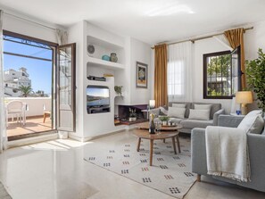 Ein modernes Wohnzimmer mit einer Couch, einem Couchtisch und einem an der Wand befestigten Fernseher. Ein Balkon mit Blick auf Gebäude und ein Sessel sind ebenfalls vorhanden.