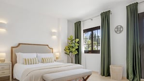 Ein Schlafzimmer mit einem Bett, Nachttischen, einer Bank, einem Fenster mit Vorhängen, einer Pflanze und einer Wanddekoration.