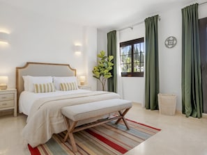 Ein Schlafzimmer mit einem Bett, Nachttischen, einer Bank, einem Fenster mit Vorhängen, einer Pflanze und einer Wanddekoration.