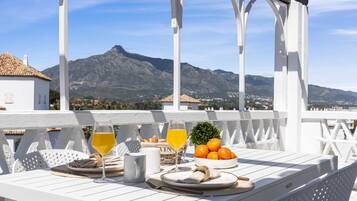 Un área de comedor exterior blanca con una mesa preparada para dos personas, con vistas a un paisaje montañoso.