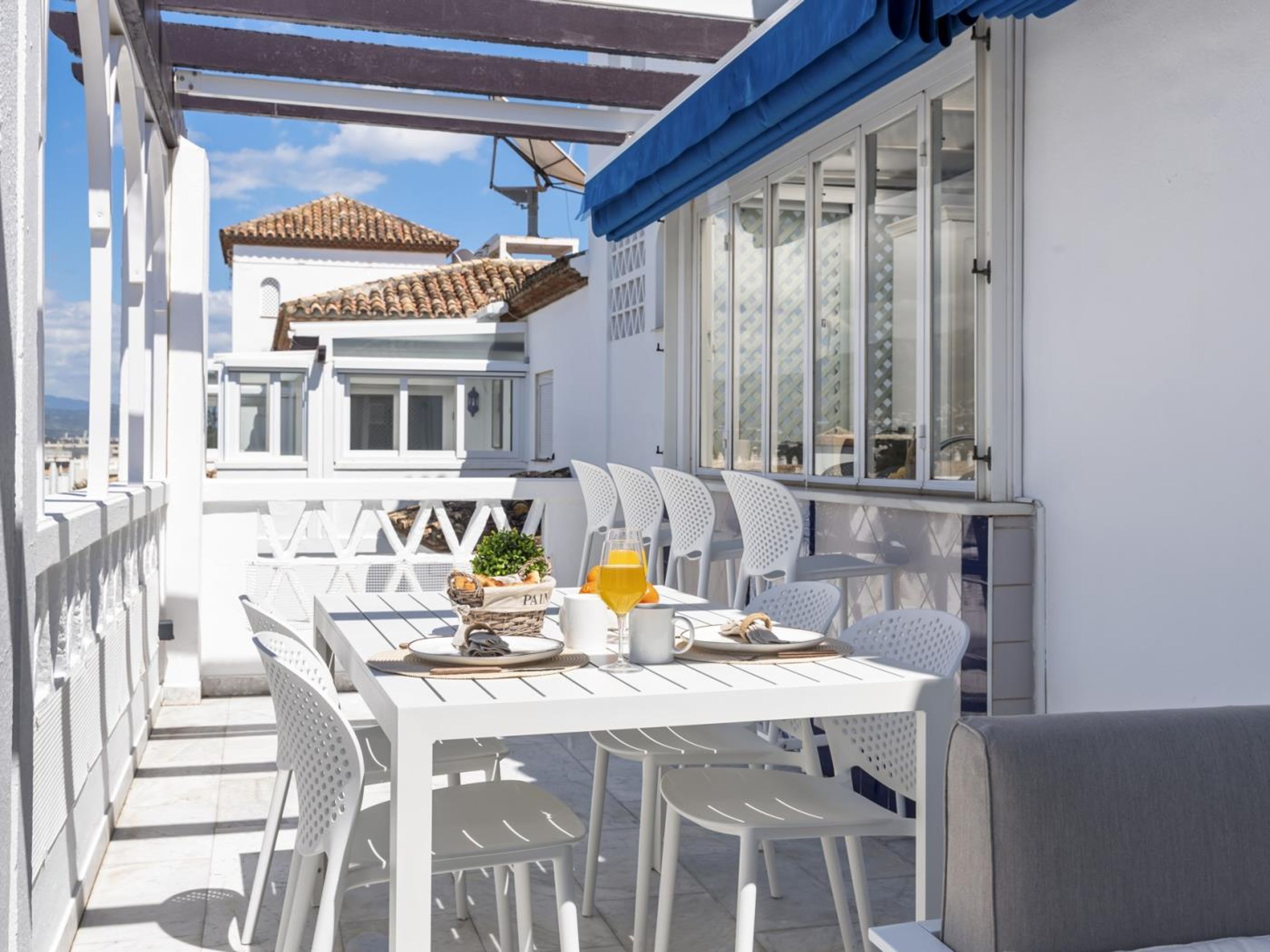 Un área de comedor exterior blanca con una mesa preparada para dos, una cesta de pan y un vaso de jugo de naranja.