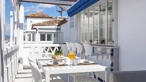 A white outdoor dining area with a table set for two, a basket of bread, and a glass of orange juice. - 01 bdr Casa Sevilla 604 (Marbella)
