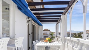 A balcony with a white table and chairs, a blue awning, and a view of buildings in the background. - 01 bdr Casa Sevilla 604 (Marbella)