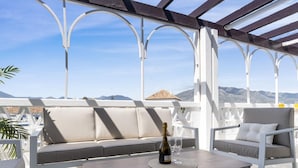 A covered outdoor seating area with a sofa and chairs, a table with a bottle and glasses, and a view of mountains. - 01 bdr Casa Sevilla 604 (Marbella)