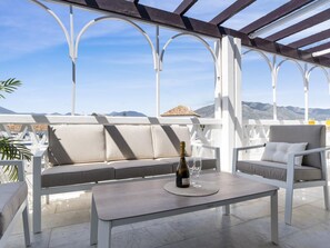 Ein überdachter Außenbereich mit einer Couch und Sesseln, einem Tisch mit Flasche und Gläsern sowie Blick auf Berge.