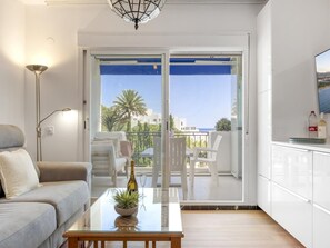 A modern living room with a sofa, a glass coffee table with a plant, a dining area with chairs, and a view of the sea through a sliding glass door. - Estudio Medina Garden 232 (Marbella)