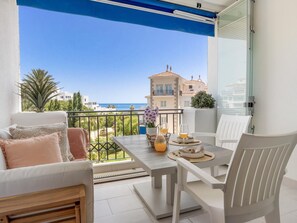 A balcony with a table set for two, a sofa, and a view of the sea and buildings. - Estudio Medina Garden 232 (Marbella)
