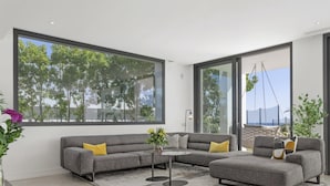 Uma sala de estar moderna com um sofá seccional cinza, uma mesa de centro e uma grande janela com vista para árvores e uma praia.