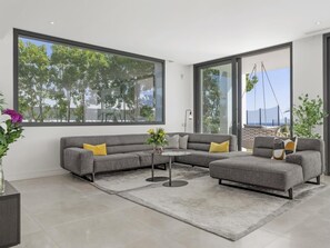 Ein modernes Wohnzimmer mit einem grauen Ecksofa, einem Couchtisch und einem großen Fenster mit Blick auf Bäume und einen Strand.