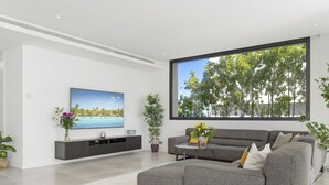 Uma sala de estar moderna com um sofá cinzento, uma televisão de tela plana e uma janela grande com vista para árvores.
