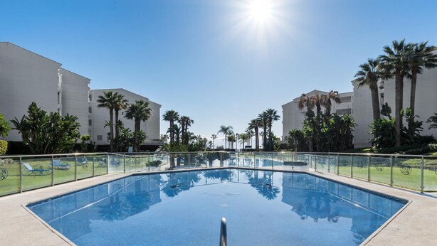 A clear blue swimming pool with a metal ladder in the center, surrounded by a low fence and palm trees. - 1 Bdr Playa Roc o 2 (Marbella)