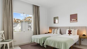 Ein Hotelzimmer mit zwei Betten, einem Sessel, einem Fenster mit Vorhängen und Bildern an der Wand.