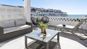 Eine Dachterrasse mit einem Korbsessel, zwei schwarzen St ühlen, einem Glastisch mit einer Flasche sowie Blick auf das Meer und Gebäude.