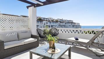 Una terraza en la azotea con un sofá de mimbre, dos sillas negras, una mesa de vidrio y una botella, con vista al mar y edificios.