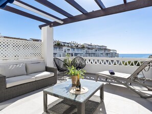 Eine Dachterrasse mit einem Korbsessel, zwei schwarzen Stühlen, einem Glastisch mit einer Flasche sowie Blick auf das Meer und Gebäude.