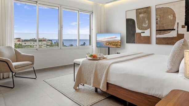 Ein modernes Schlafzimmer mit einem großen Bett, einem Sessel, einem Fernseher und Meerblick.
