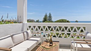 Ein Balkon mit weißer Gartenmöbeln, einem Holztisch mit Flasche und Glas sowie Blick auf das Meer und Grünflächen.