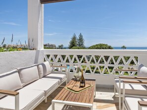 A balcony with white outdoor furniture, a wooden table with a bottle and glass, and a view of the sea and greenery. - Casa Granada 2 44 in Puerto Ban s - Marbella (Marbella)