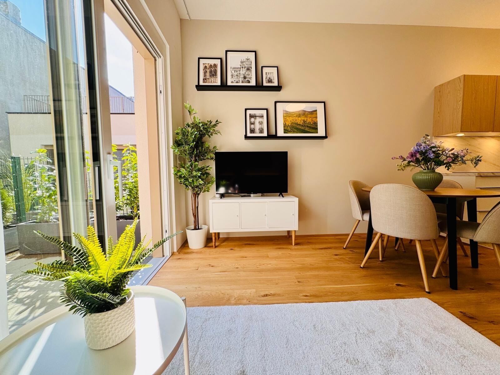 A modern living room with a flat-screen TV, a dining area with chairs, and a large window offering a view of greenery.